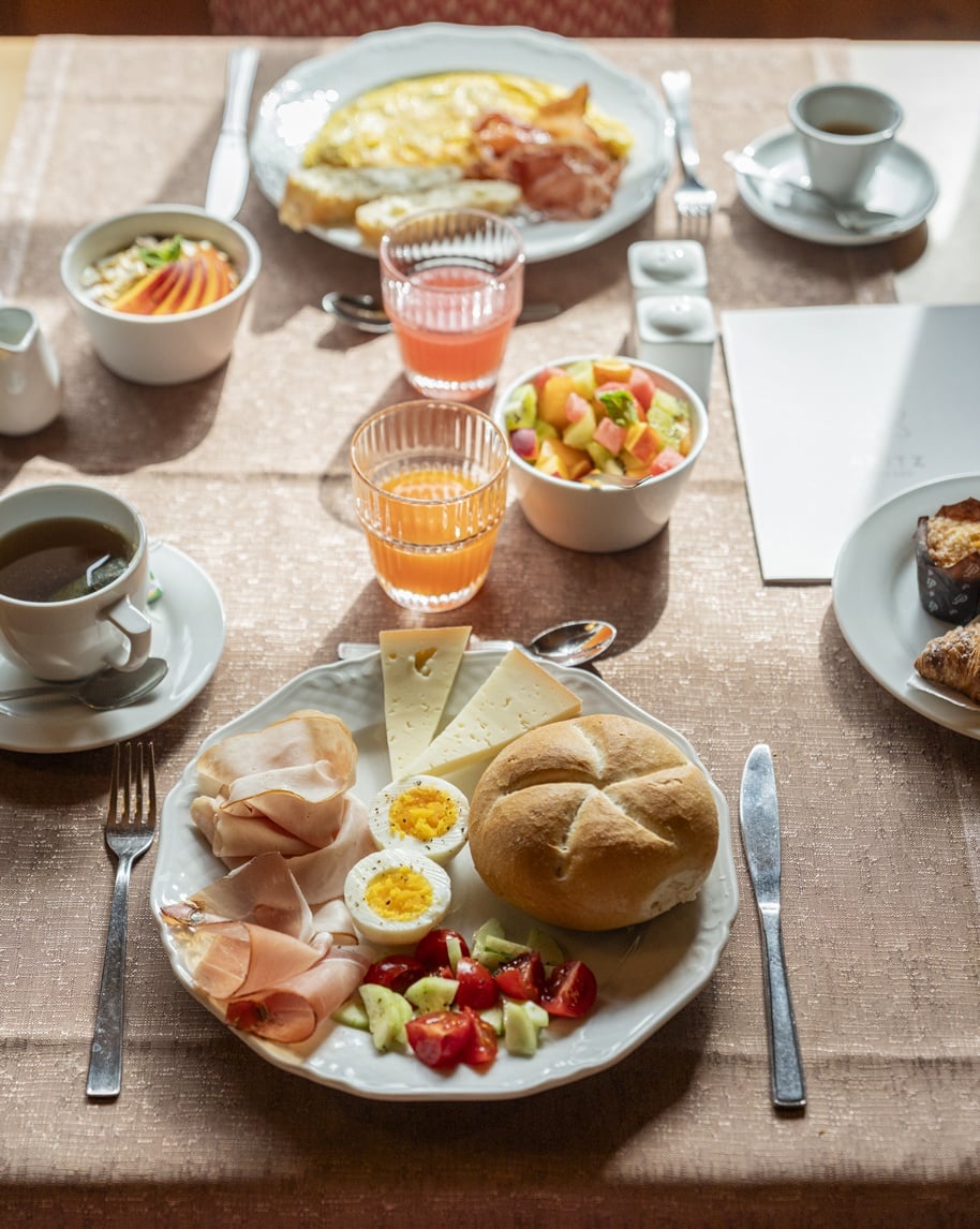 Tavola da colazione con piatto: panino, formaggi, affettati, uova sode e insalata; bicchieri di succo e caffè