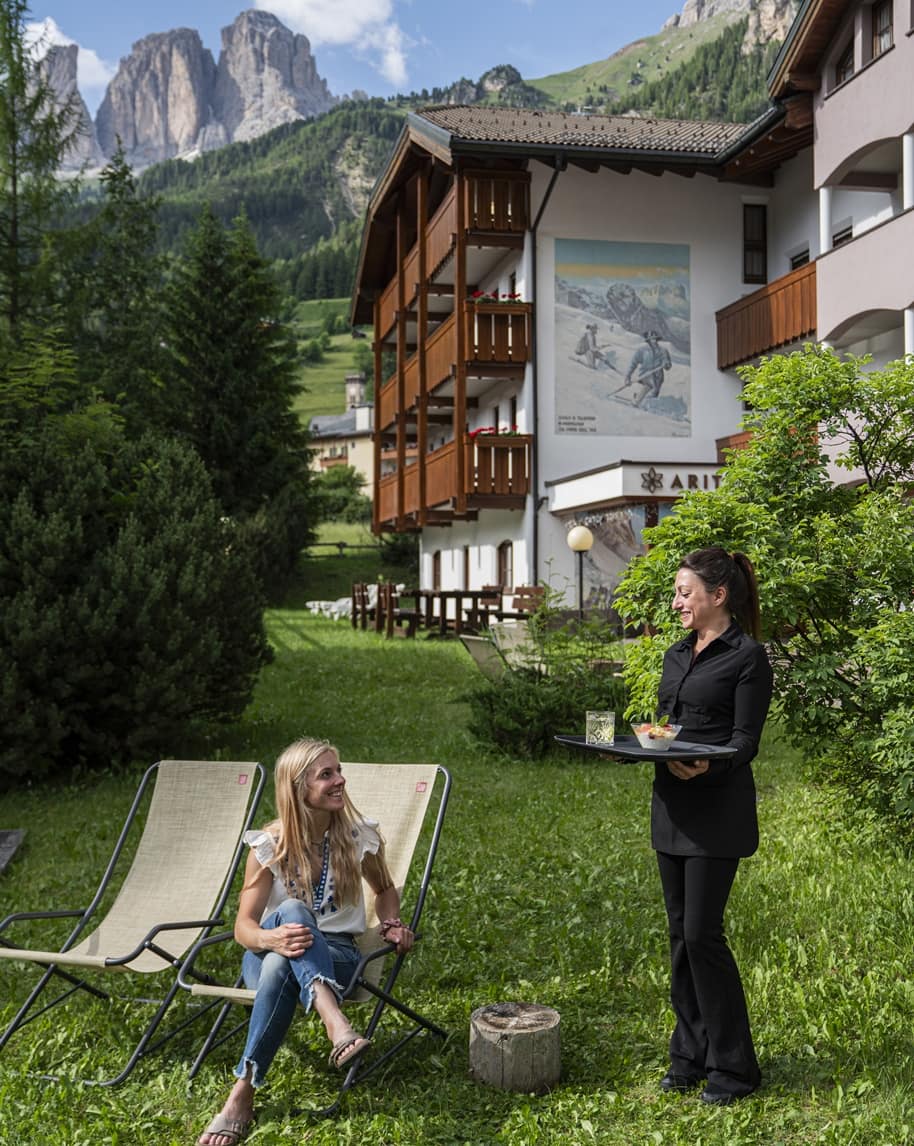 Donna su sdraio sorride mentre una cameriera le porta un vassoio nel giardino di un Hotel montano con balconi in legno