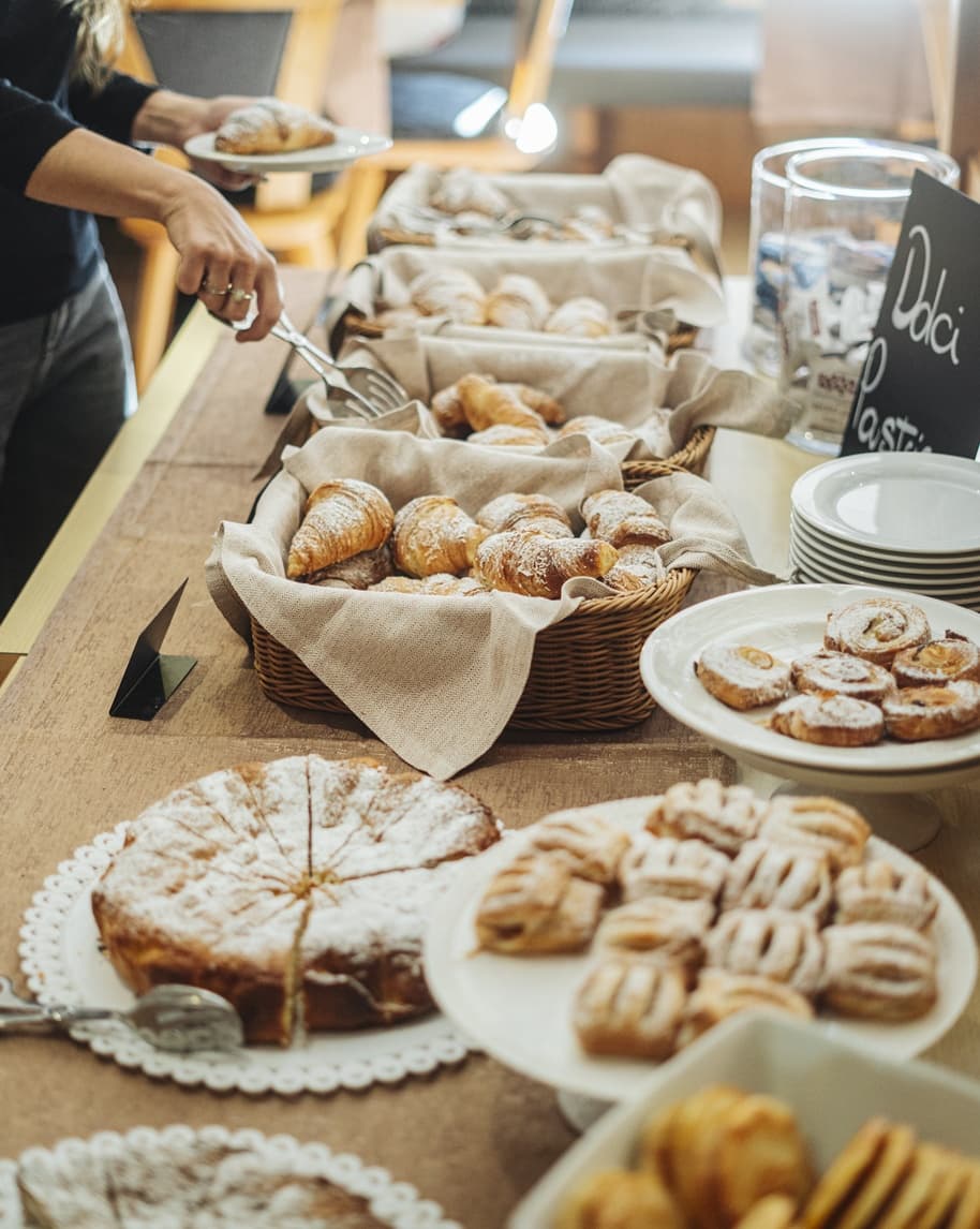 Cesti e piatti di cornetti e pasticcini su buffet con cartello 'Dolci'; persona prende un cornetto con pinza