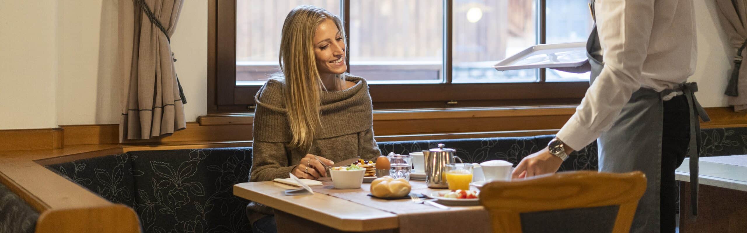 Donna seduta a tavolo mentre un cameriere serve la colazione: brioche, succo, uovo sodo, tazza e teiera
