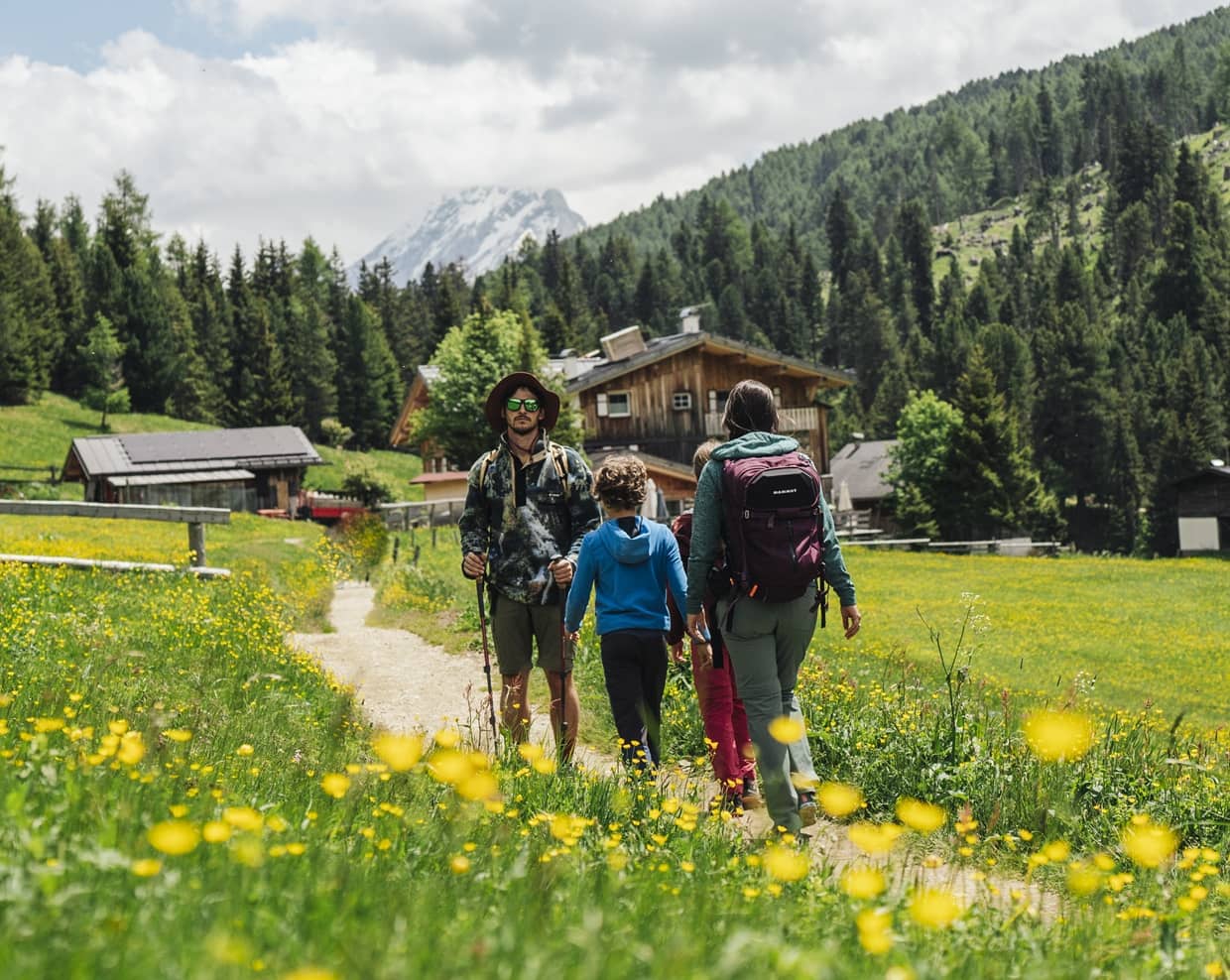 Quattro escursionisti su sentiero fiorito verso baite in un prato alpino con foresta e montagna sullo sfondo
