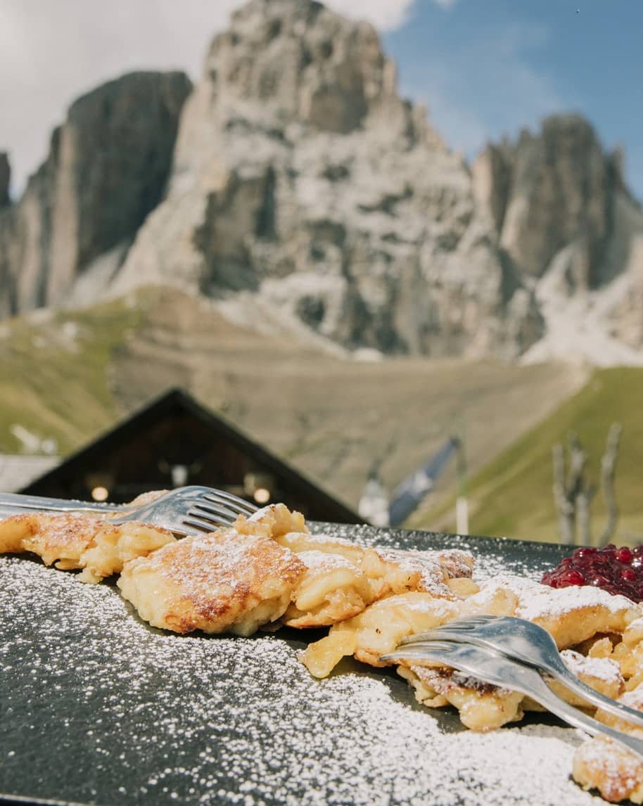 Frittelle con zucchero a velo e marmellata su piatto con forchette, montagne sullo sfondo