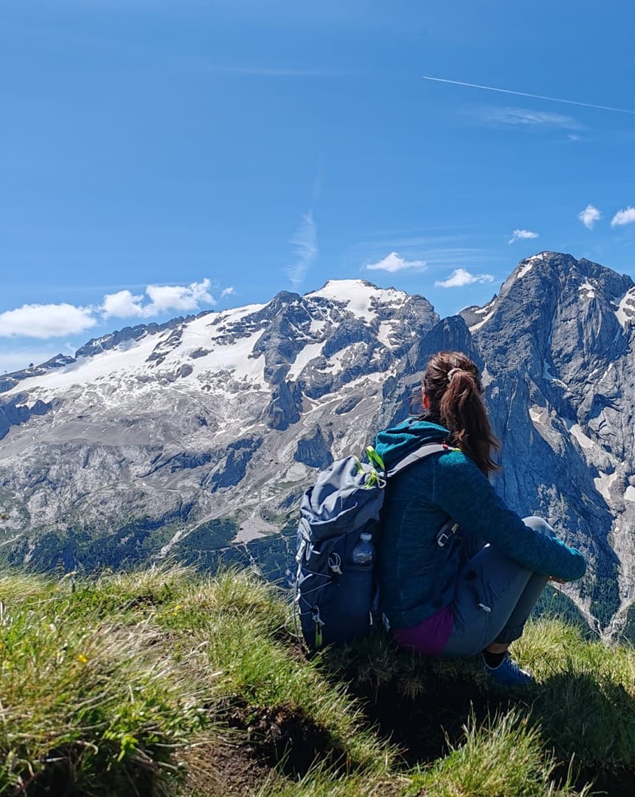 Escursionista seduta di spalle su prato guarda catena montuosa con vette innevate e pareti rocciose