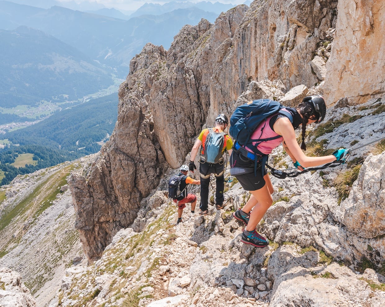 Tre escursionisti in casco e imbraghi agganciati a cavo risalgono una ferrata su parete rocciosa sopra una vallata