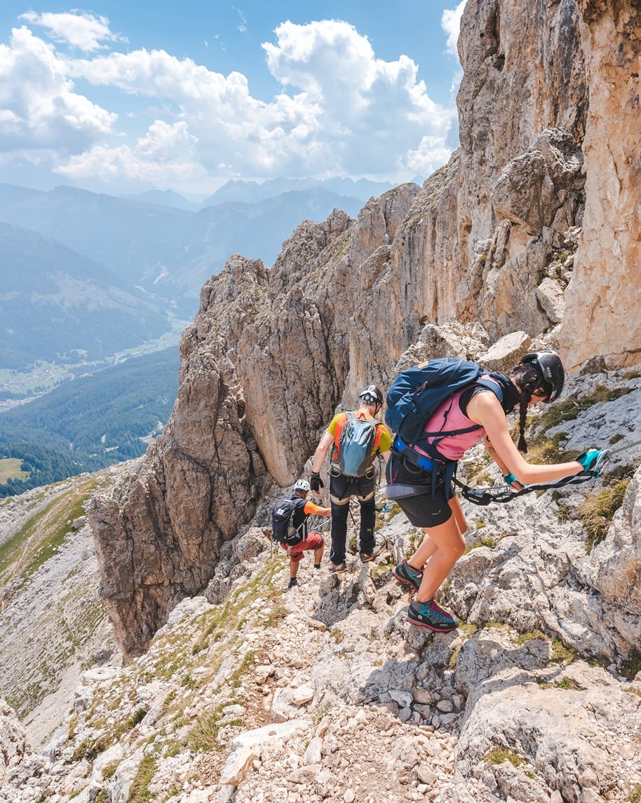 Tre escursionisti assicurati su cengia rocciosa mentre scendono lungo una parete montana, valle sottostante