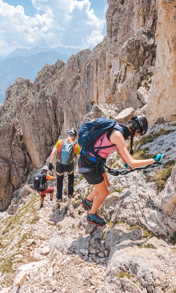 Tre escursionisti con casco e zaini calandosi lungo una parete rocciosa alpina tenendosi a una fune metallica