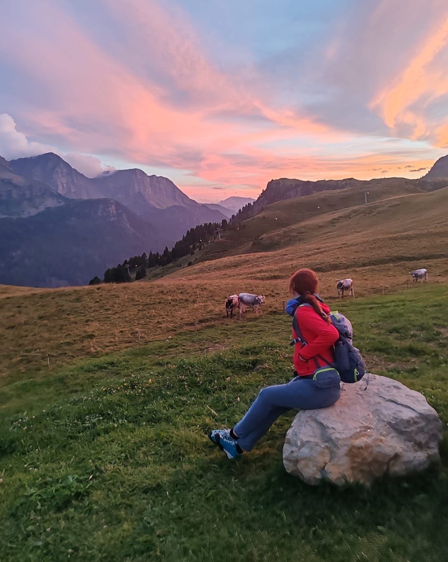 Persona seduta su un masso con zaino guarda pascolo con mucche e montagne al tramonto