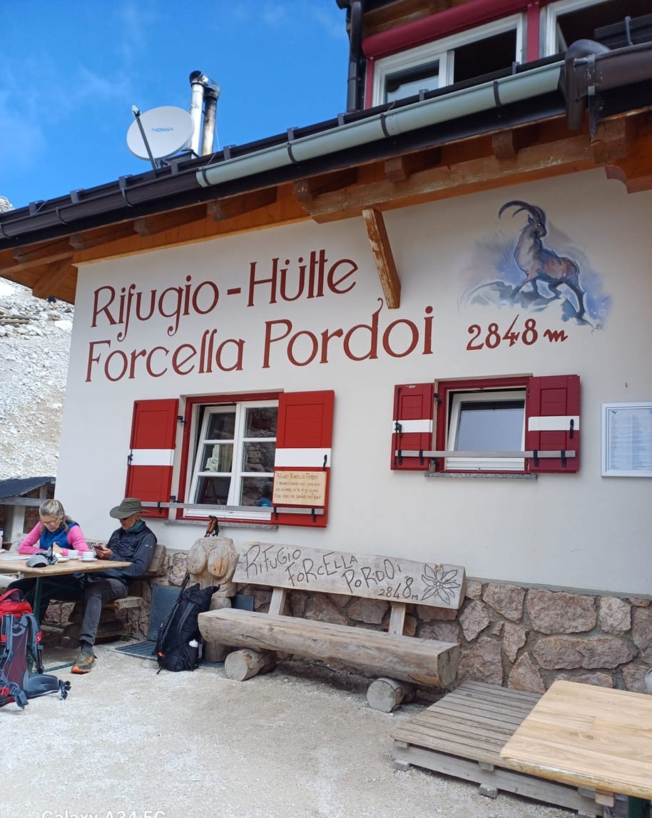 Rifugio Forcella Pordoi 2848 m: edificio alpino con persiane rosse, panchina in legno e due escursionisti seduti