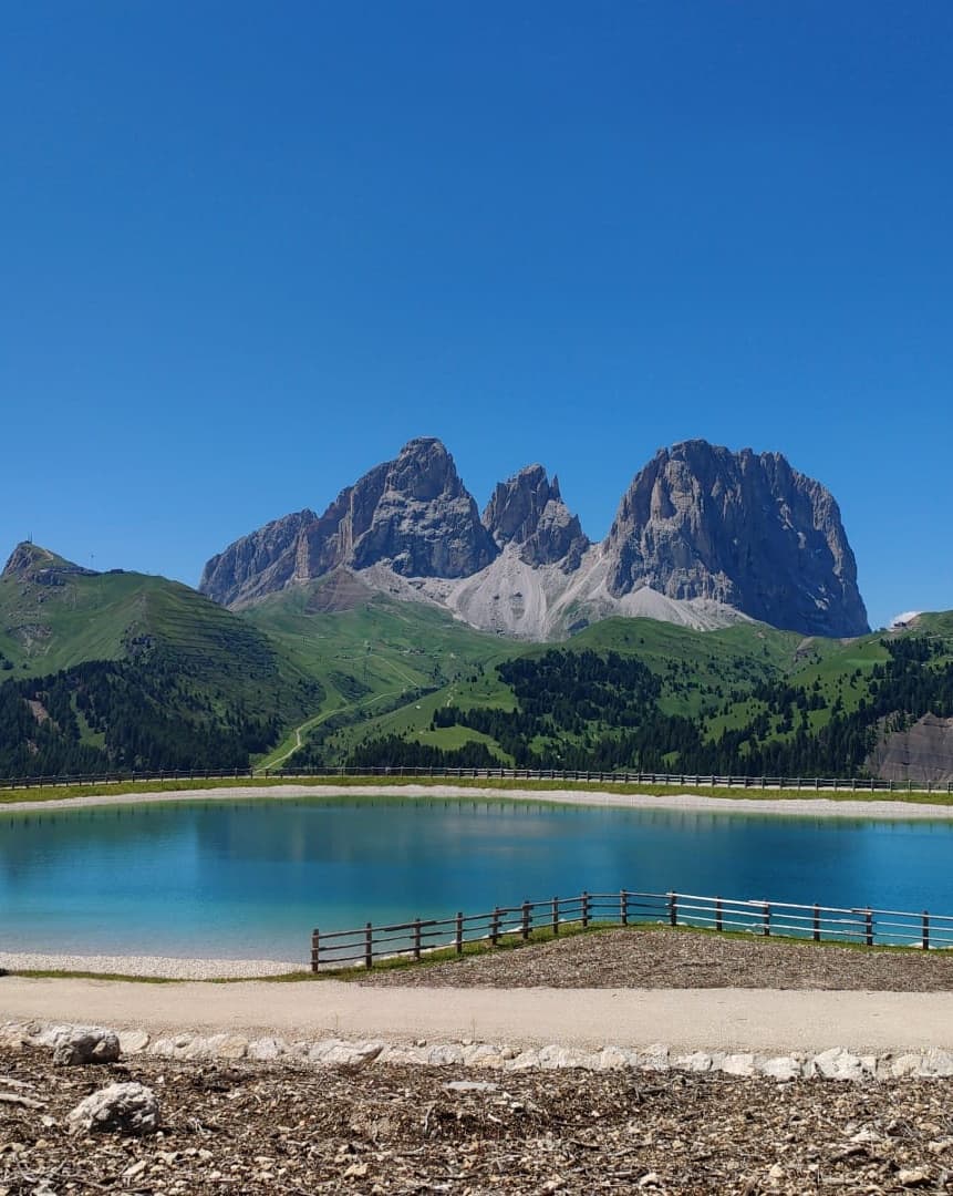 Specchio d'acqua turchese davanti a recinzione in legno con colline verdi e creste rocciose sullo sfondo