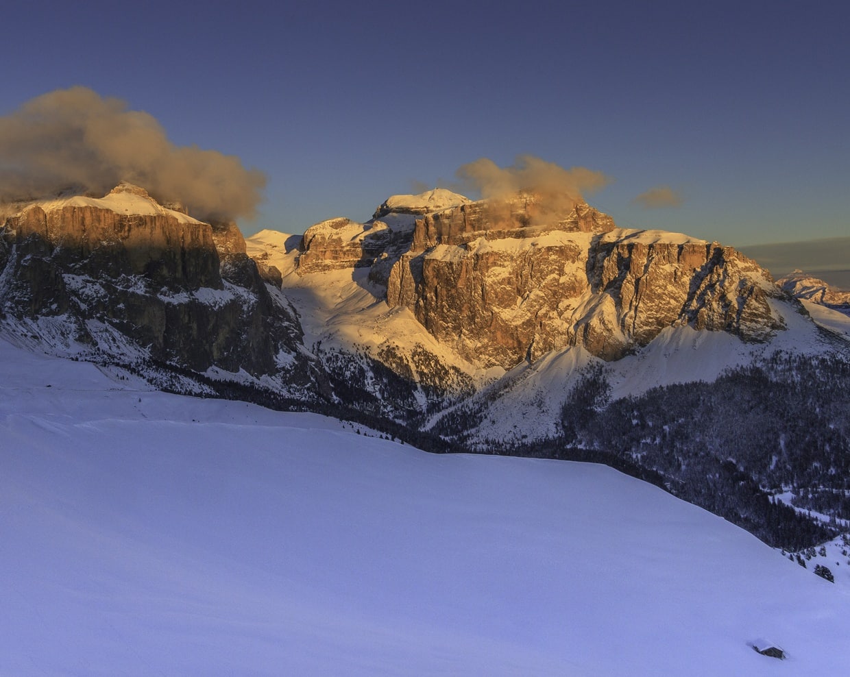 Catena di montagne rocciose innevate con nuvole basse, cime illuminate e ampia distesa di neve in primo piano