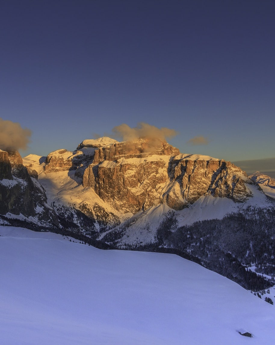 Altopiano innevato in primo piano e pareti rocciose delle Dolomiti al tramonto con nuvole basse