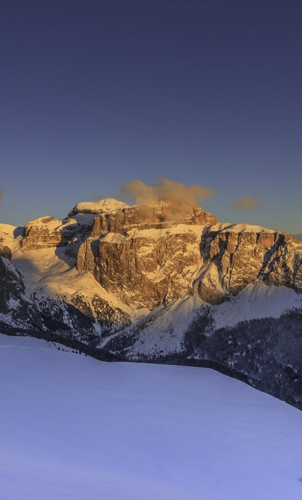 Massiccio roccioso innevato illuminato dal sole basso, nube sul crinale e ampio pendio di neve in primo piano