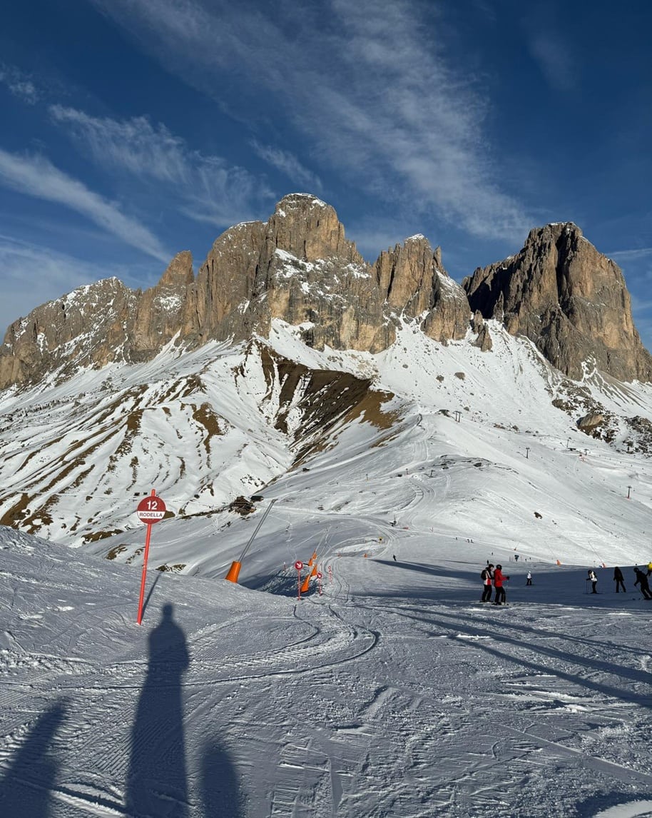 Montagna rocciosa con neve, pista da sci in primo piano, cartello 12 RODELLA e sciatori