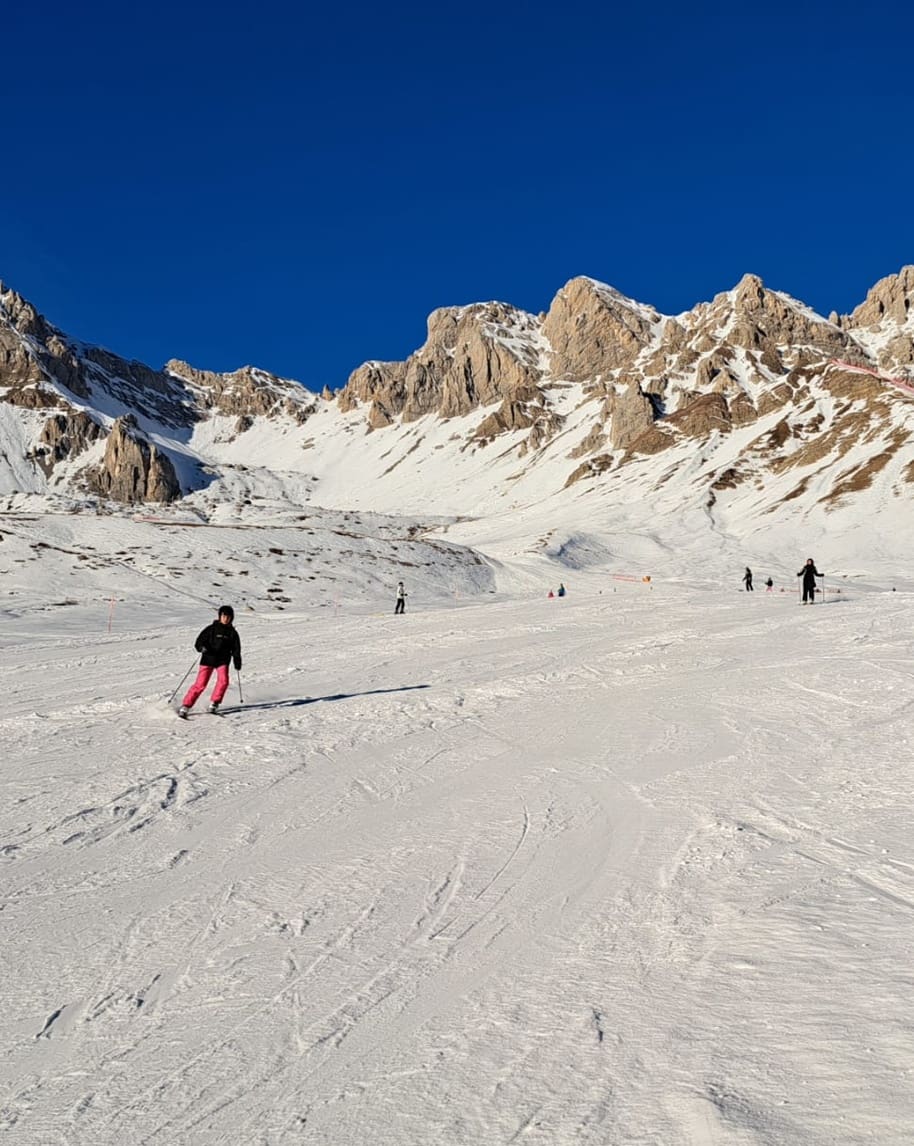 Persona sugli sci in pantaloni rosa scende su ampio pendio innevato davanti a cime rocciose e cielo azzurro