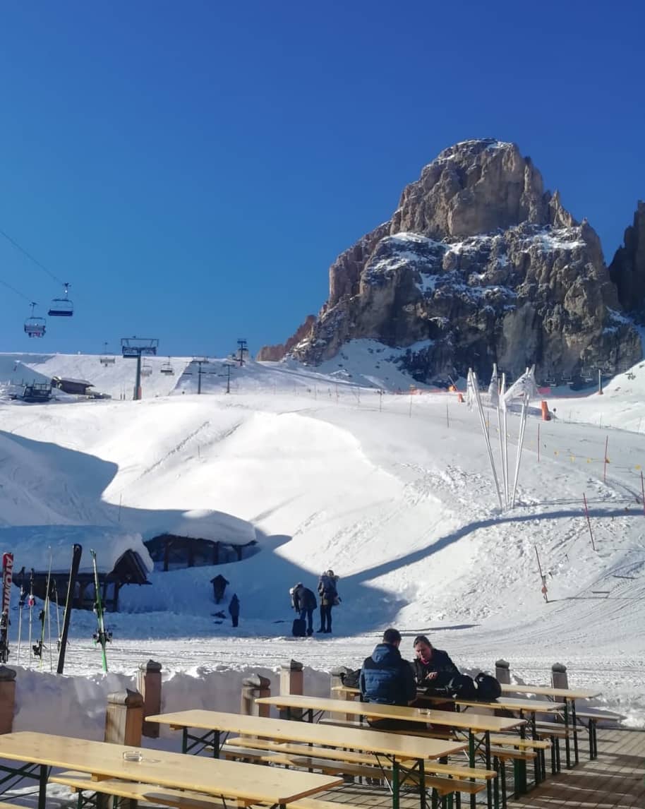 Cima rocciosa innevata sullo sfondo, pista da sci con seggiovie e tavoli esterni con due persone sedute