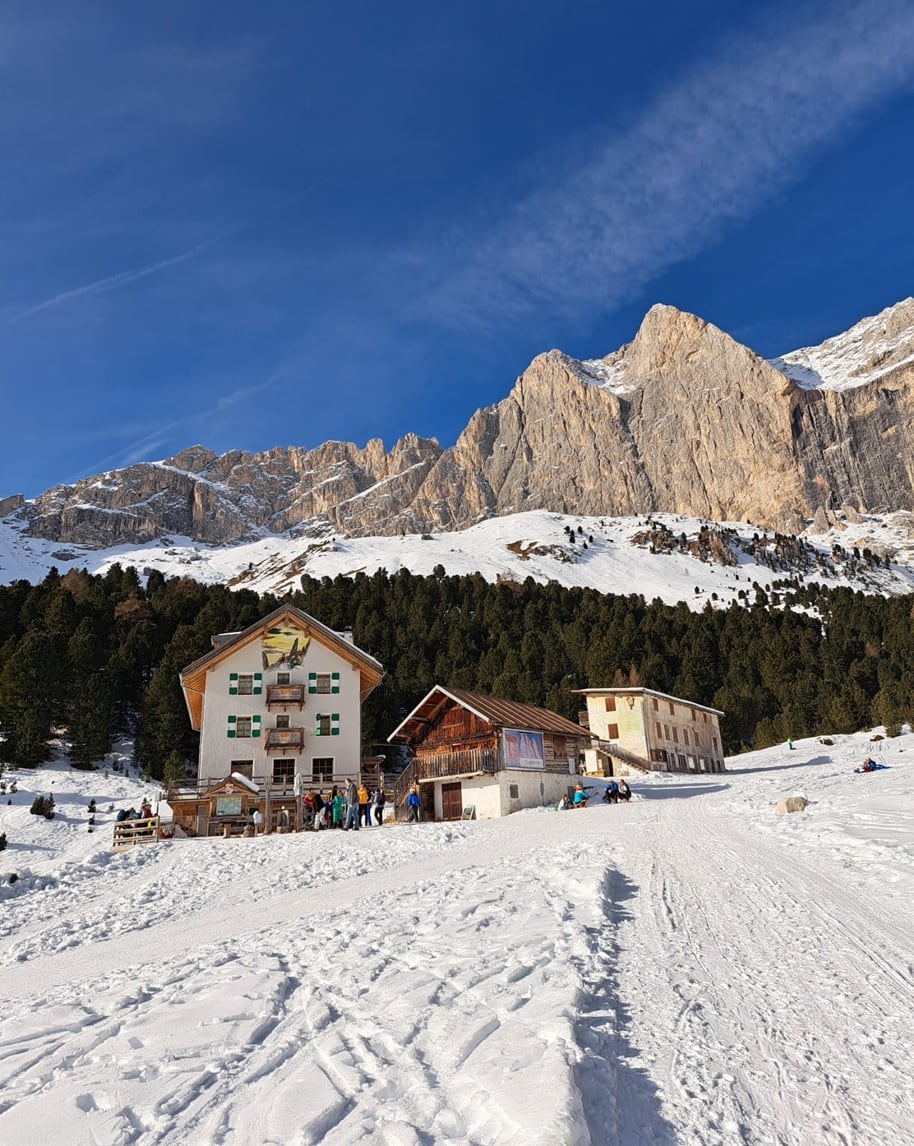 Tre edifici alpini con persone davanti su neve, bosco di pini e alte pareti rocciose innevate sullo sfondo
