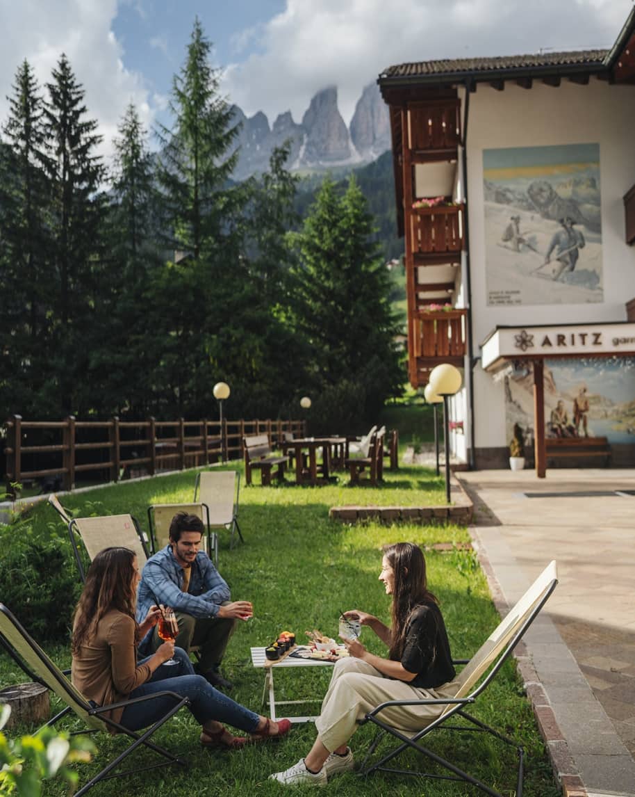 Tre persone sedute su sdraio in giardino che condividono drink e stuzzichini davanti all'Hotel Aritz e monti.