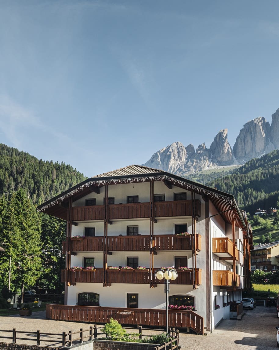 Hotel a tre piani con balconi in legno e fioriere, foresta e picchi rocciosi sullo sfondo.