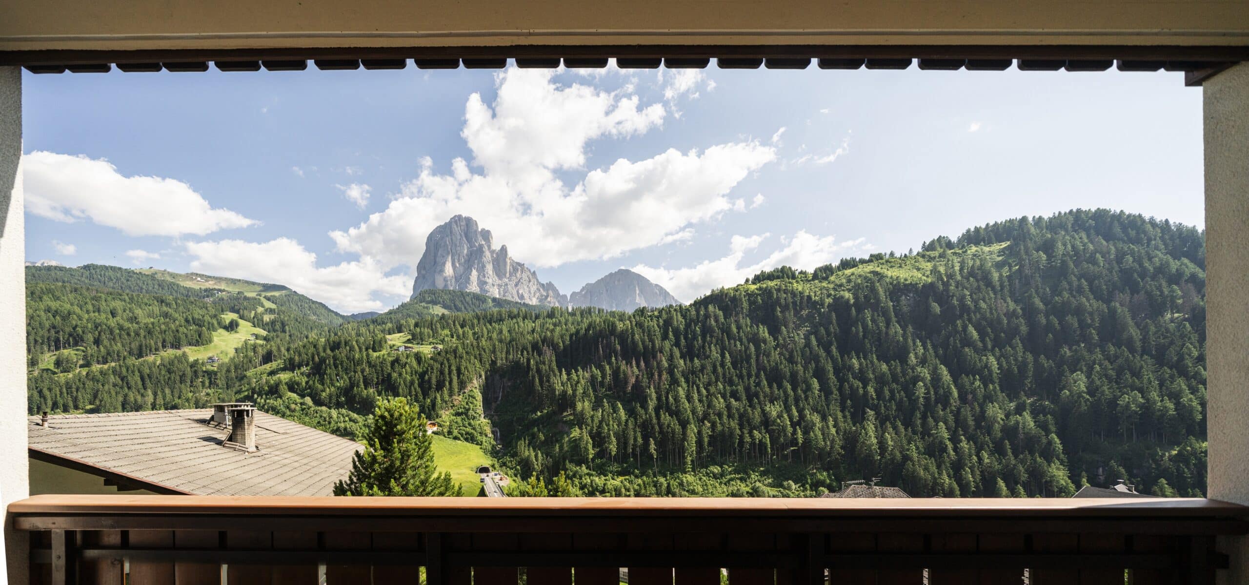Ringhiera di balcone in primo piano, vallata boscosa con colline e parete rocciosa prominente sullo sfondo