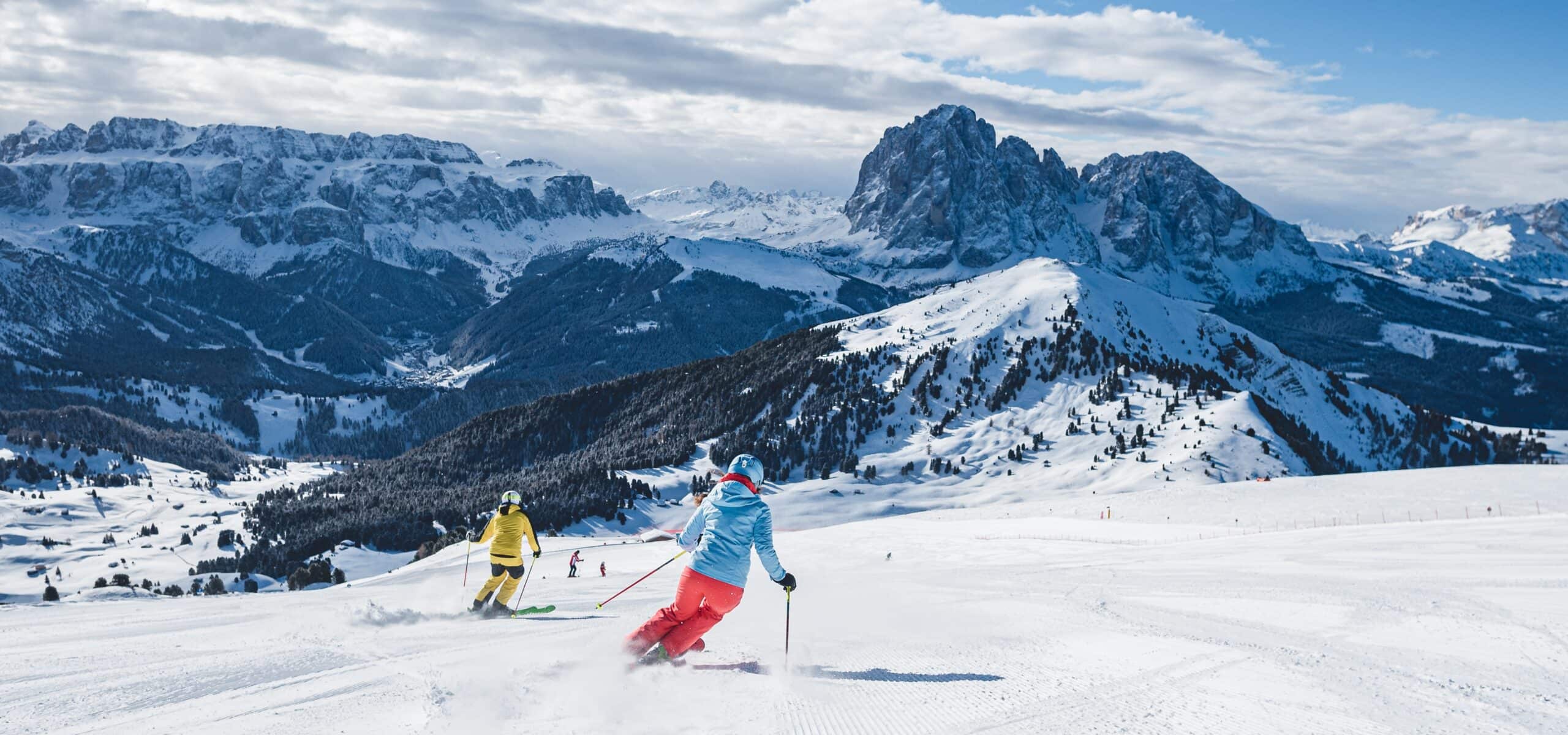 Due sciatori in giacche colorate scendono una pista innevata con alte montagne rocciose sullo sfondo