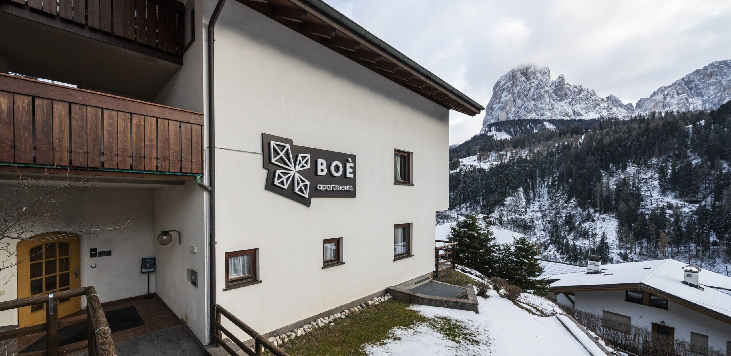 Edificio Boè apartments con balcone e ingresso; montagna innevata e bosco sullo sfondo