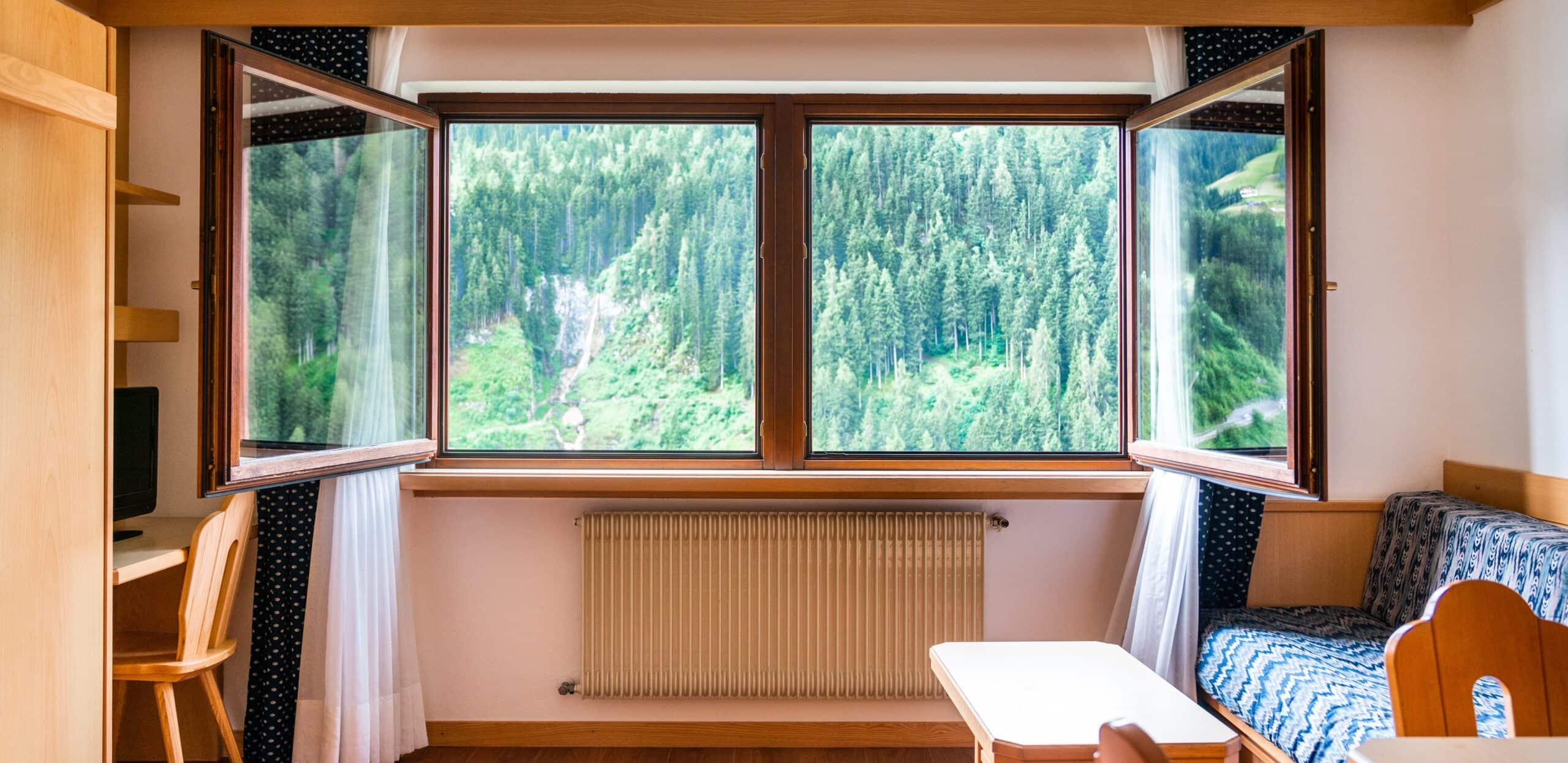 Finestra aperta su bosco e montagna vista da stanza con radiatore sotto e mobili in legno.