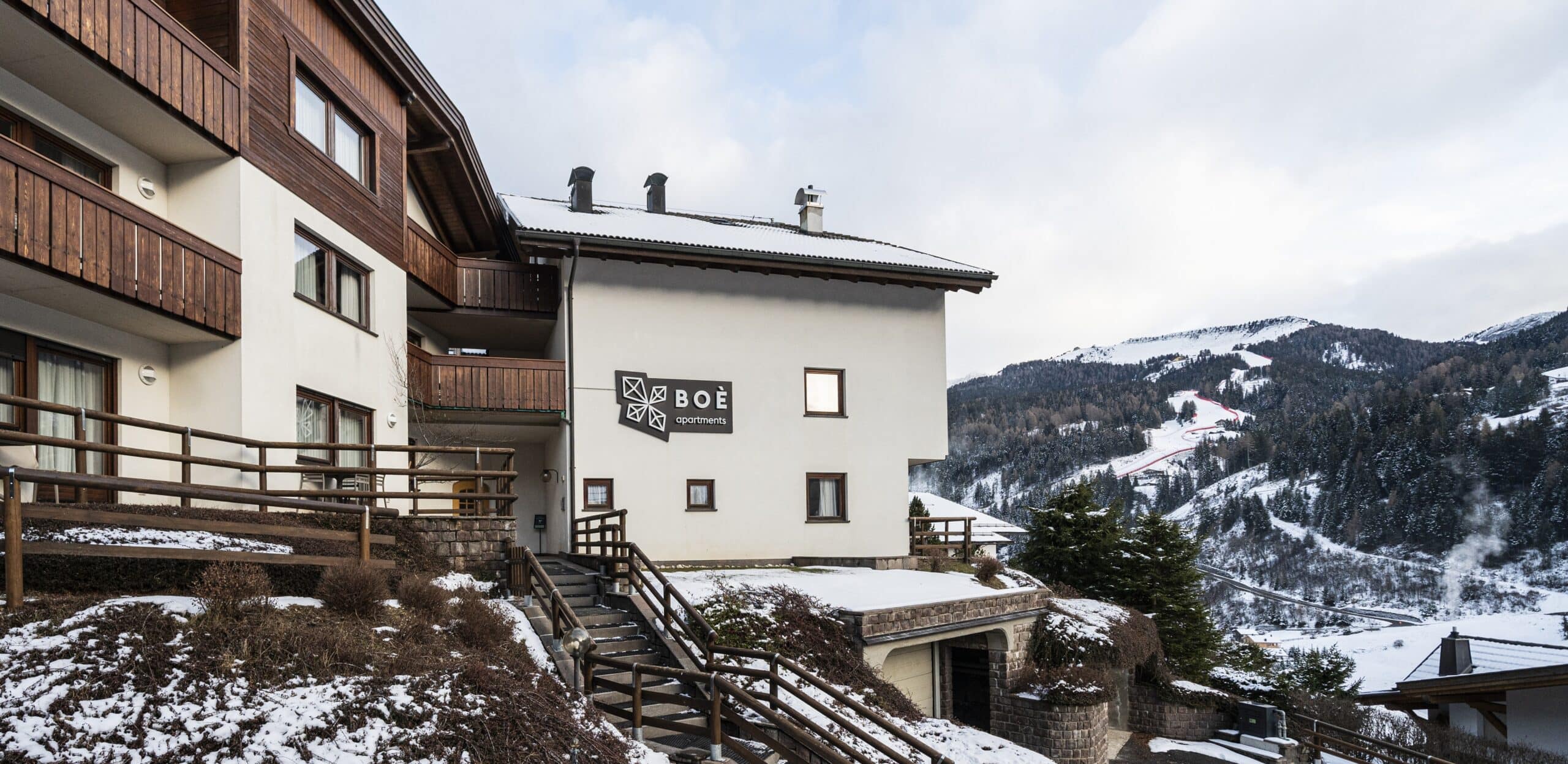 Edificio BOÈ apartments su collina innevata con balconi in legno e montagne con piste da sci sullo sfondo