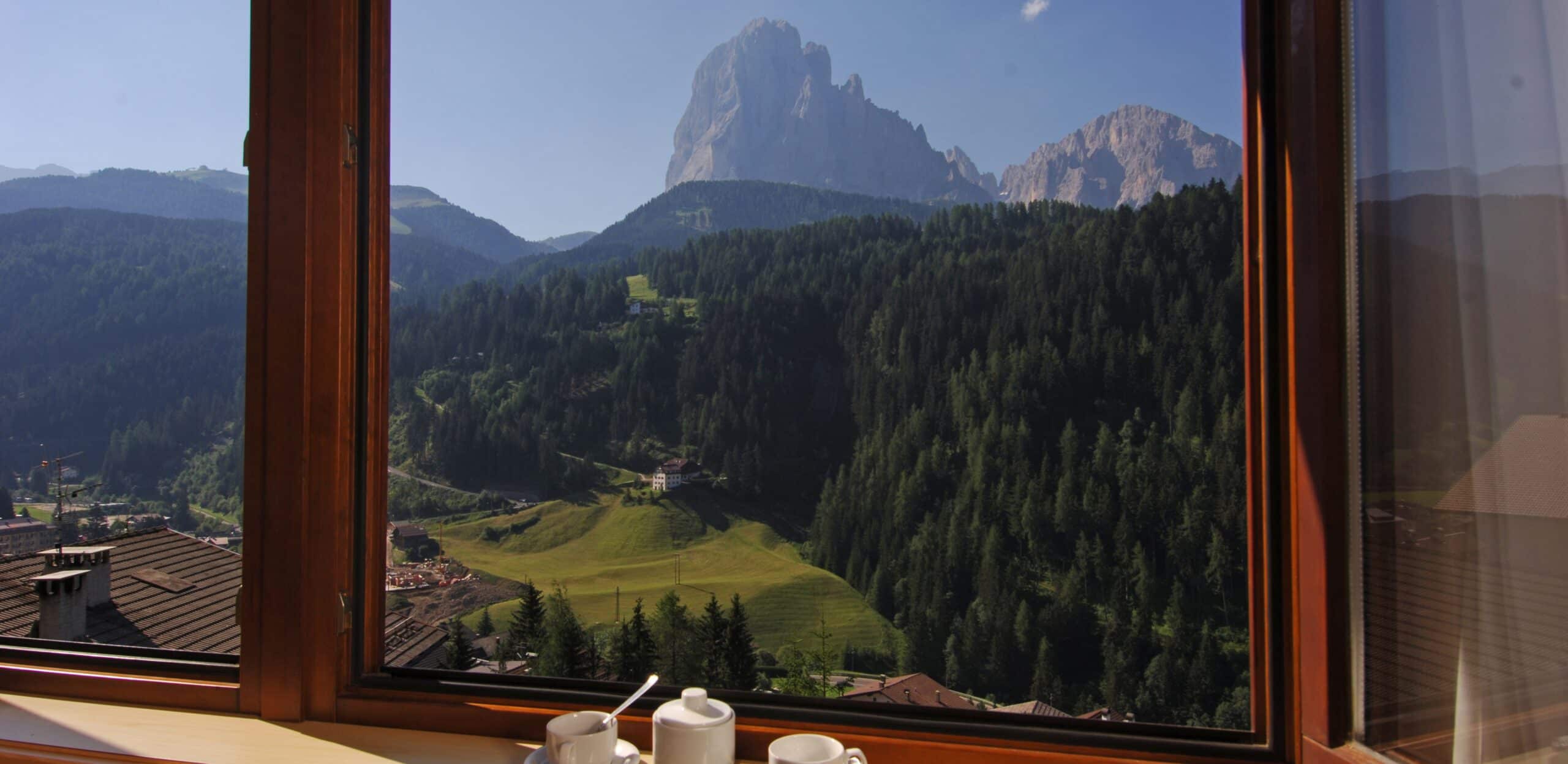 Finestra aperta su vallata montana con cime rocciose, boschi e tazze con teiera sul davanzale