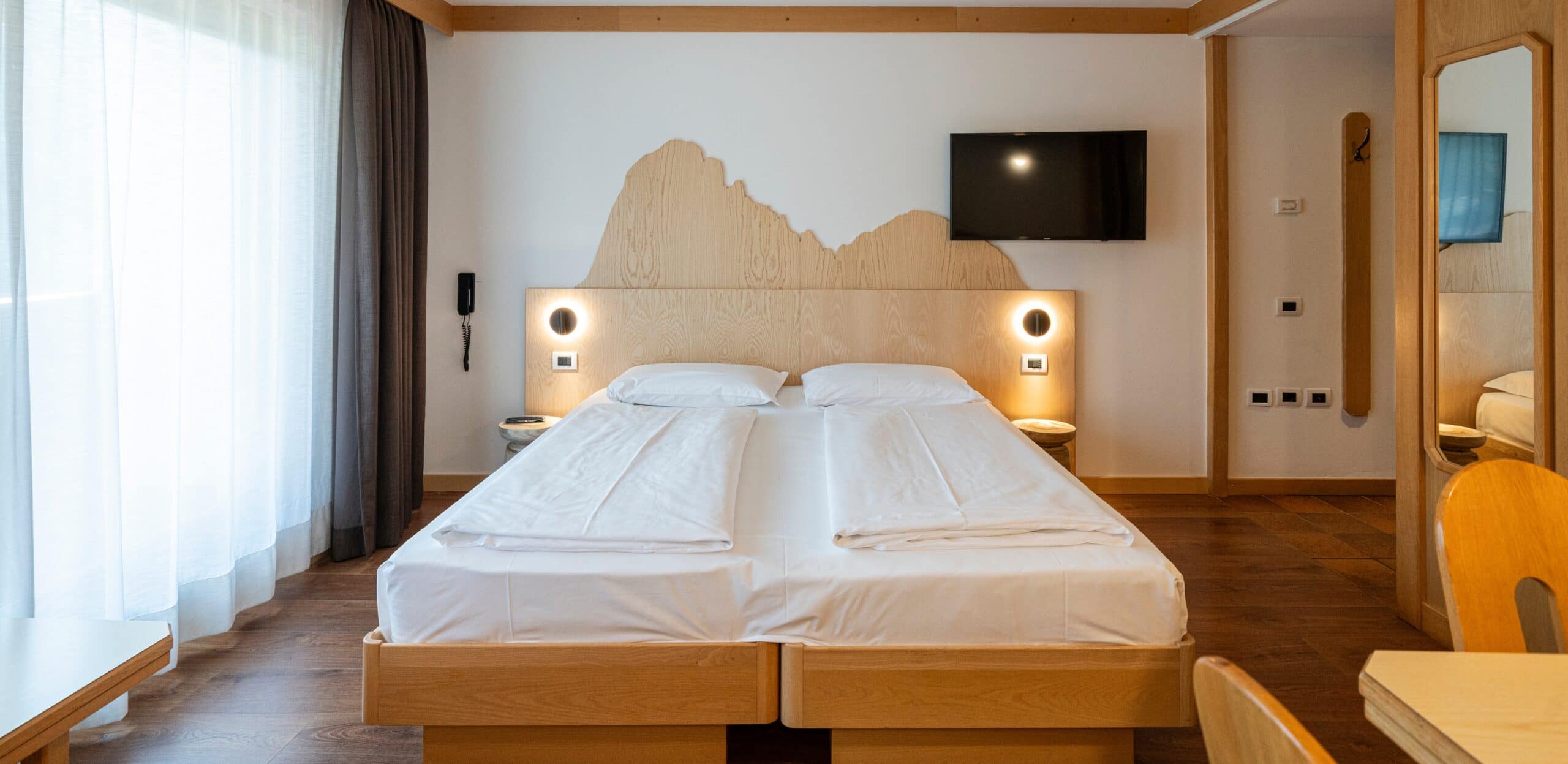 Letto matrimoniale con testiera sagomata in legno, lampade circolari, TV a parete e finestra con tende