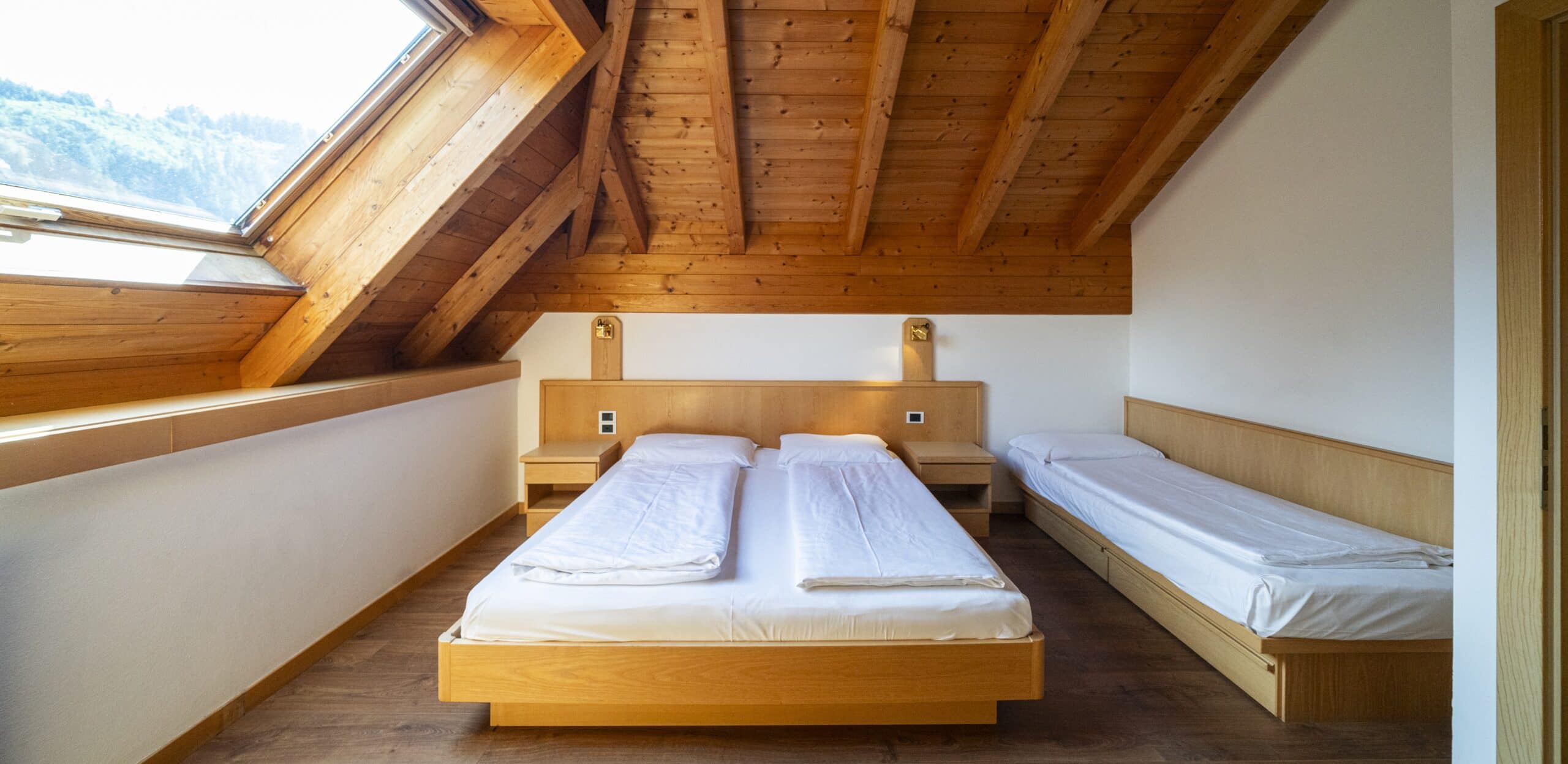 Mansarda con travi in legno, finestra a tetto, letto matrimoniale centrale e letto singolo laterale.