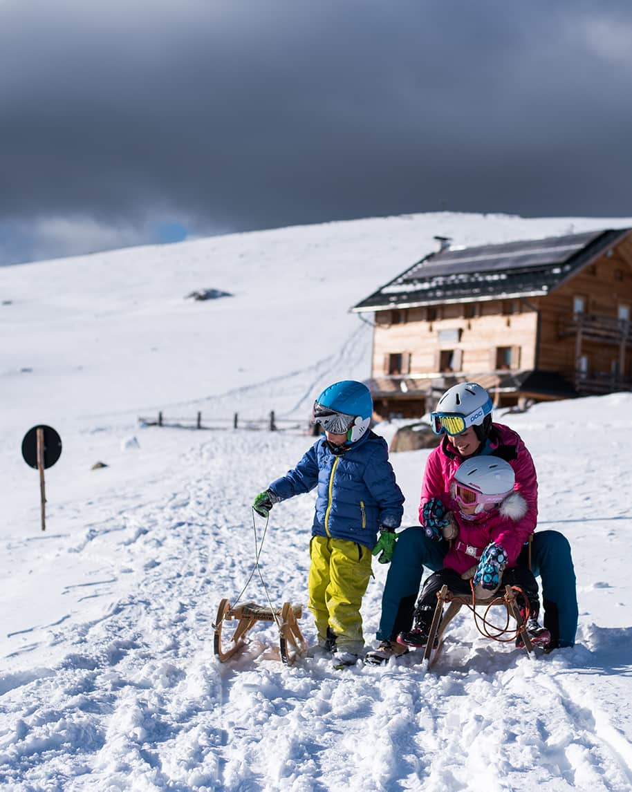 Donna con due bambini su slitte nella neve davanti a una baita sotto un cielo nuvoloso