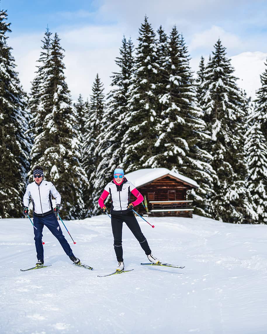 Due sciatori di fondo su pista davanti a una baita e abeti innevati