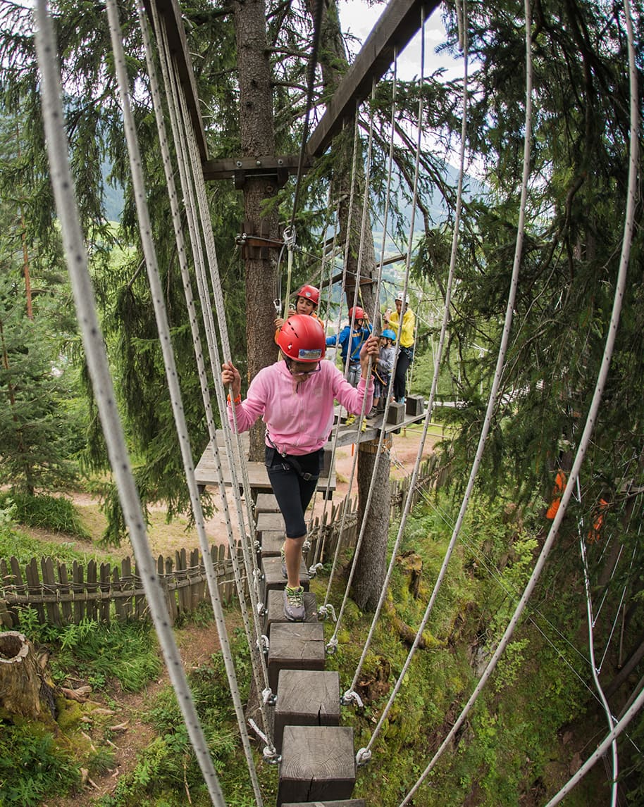 Persona con casco rosso e giacca rosa attraversa passerella sospesa in un percorso tra gli alberi, altri dietro