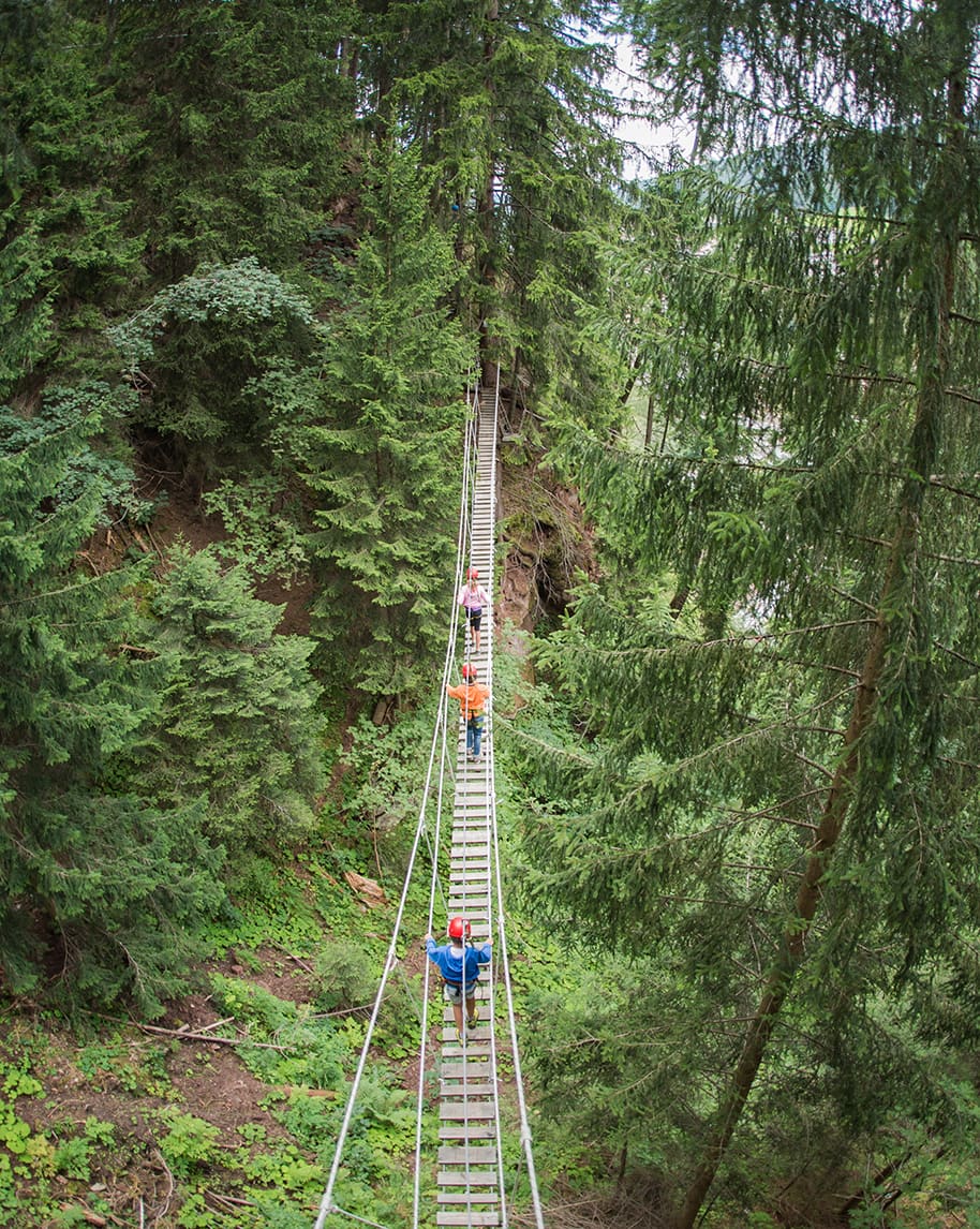 Tre persone con caschi attraversano un ponte sospeso a pioli tra gli alberi di una foresta