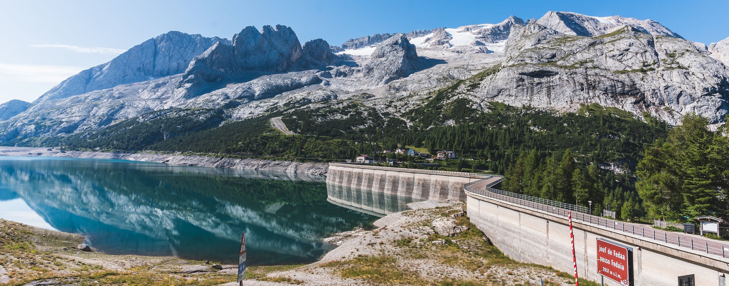 Staumauer am türkisfarbenen Bergsee, spiegelnde Gipfel, kleine Häuser am Ufer; Schild Passo Fedaia.