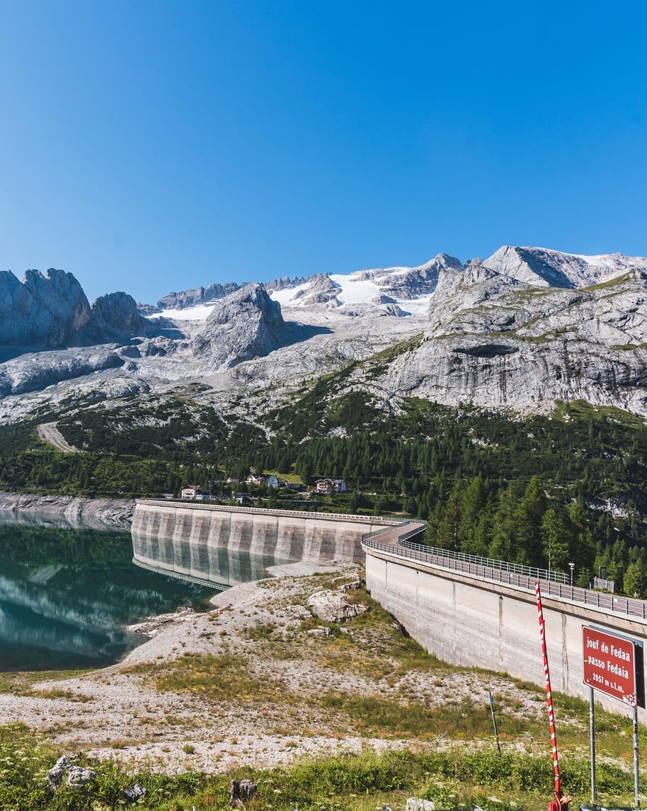 Diga curva su lago che riflette montagne rocciose e ghiacciaio, cielo azzurro; cartello Passo Fedaia