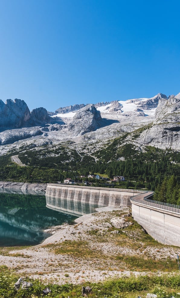 Diga curva su lago alpino, riflesso sull'acqua, montagne rocciose con ghiacciaio e case boschive