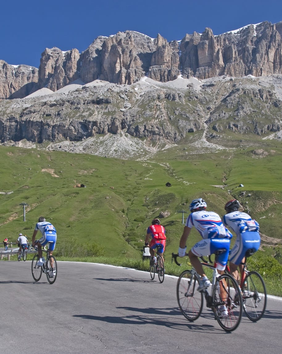 Gruppo di ciclisti in salita su strada di montagna con prati verdi e pareti rocciose sullo sfondo