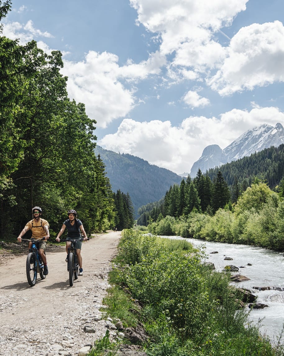 Due ciclisti su sentiero sterrato accanto a un fiume, boschi e montagne innevate sullo sfondo