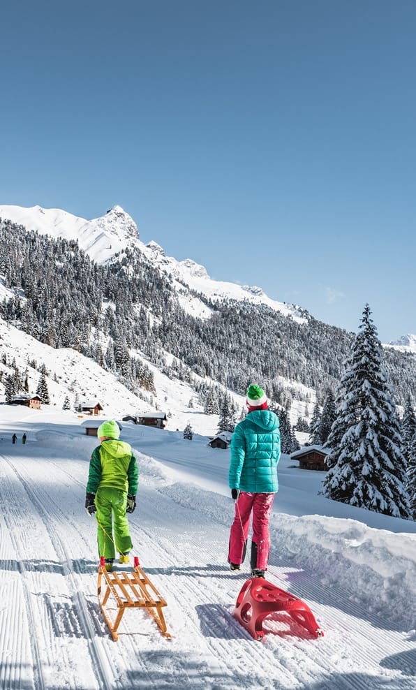 Due persone di schiena che trascinano slittini su pista innevata verso baite e montagne