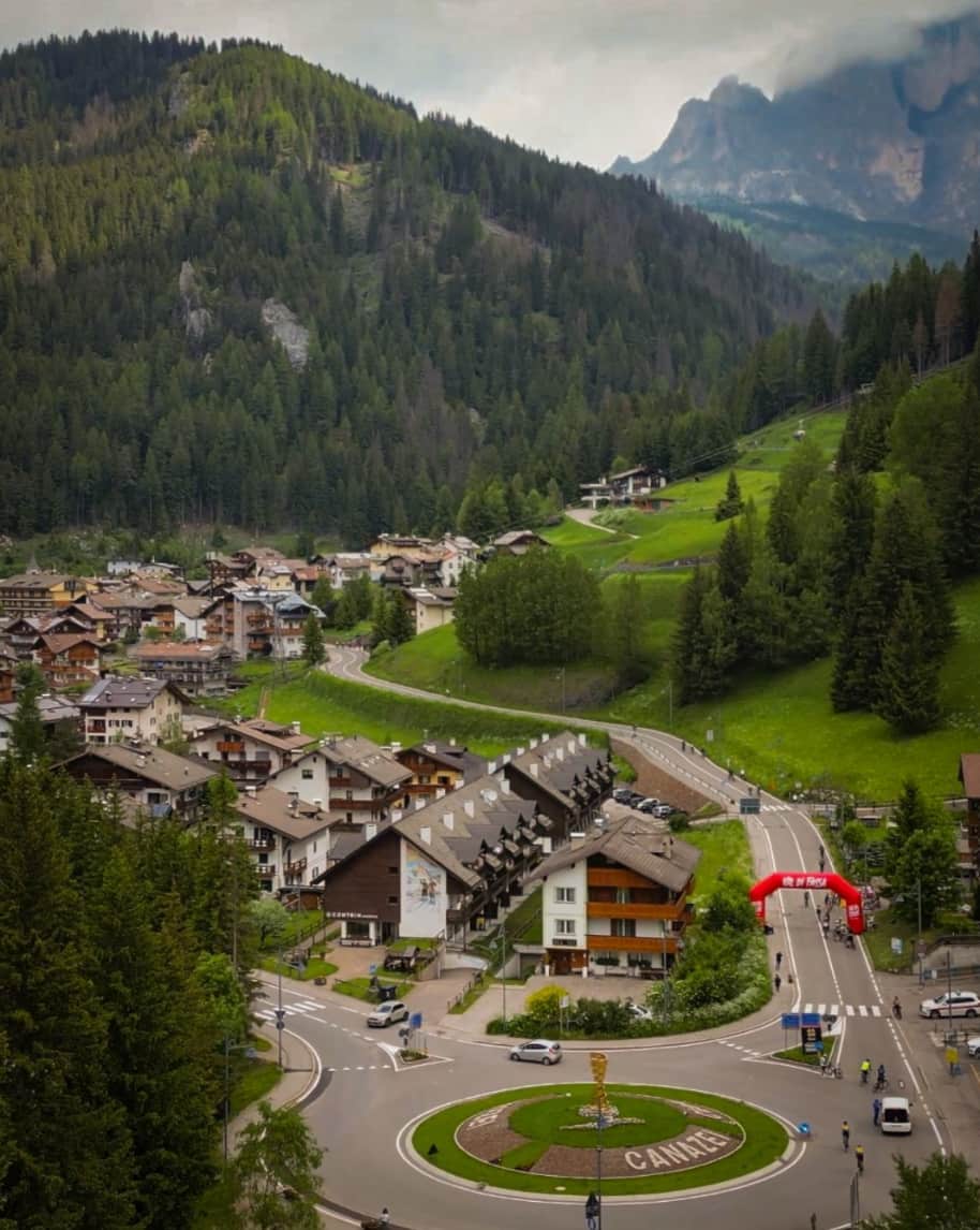 Paese alpino con case in legno, rotatoria con scritto CANAZEI, strada che sale tra prati e boschi