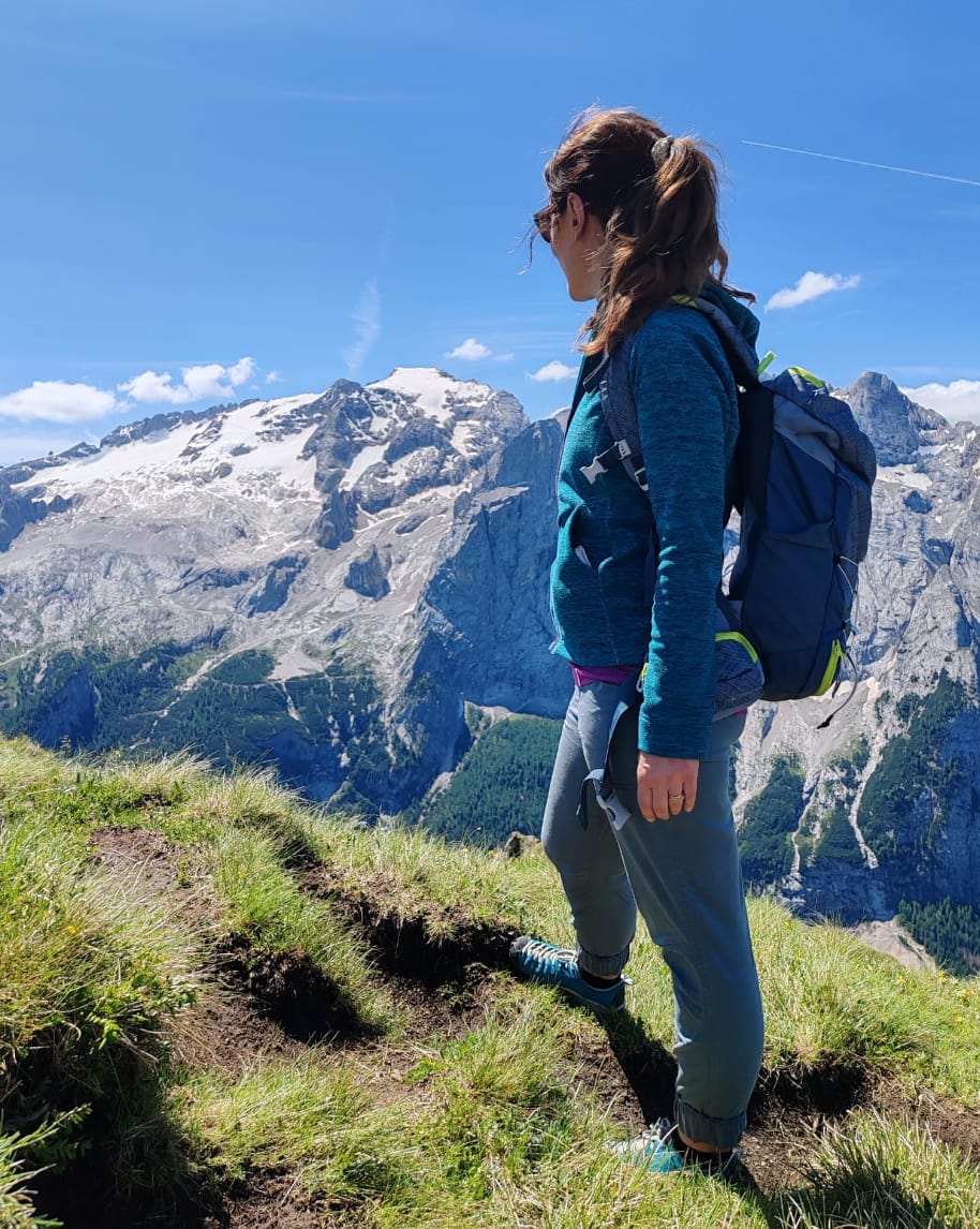 Donna con zaino in piedi su cresta erbosa che guarda una catena montuosa innevata
