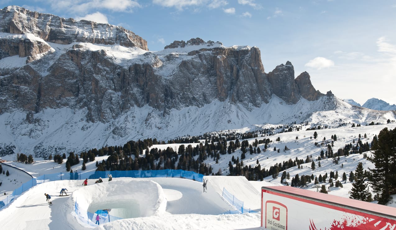 Pareti rocciose innevate sullo sfondo; in primo piano parco neve con halfpipe e snowboarder