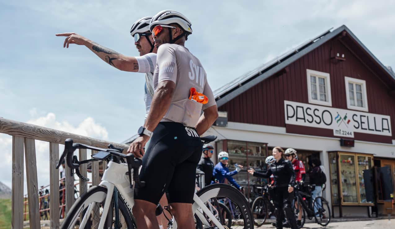 Due ciclisti con bici, uno indica verso sinistra davanti alledificio con insegna PASSO SELLA mt 2244.