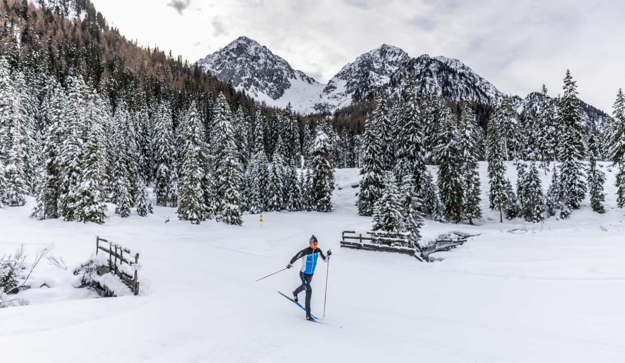 Sciatore di fondo scivola su pista innevata davanti a abeti coperti di neve e due cime montuose sullo sfondo