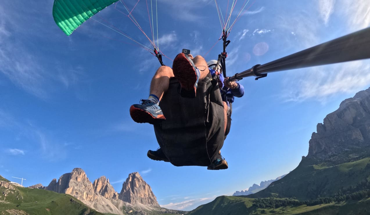 Persona in parapendio biposto vista dal basso, gambe a penzoloni e paesaggio montano sottostante
