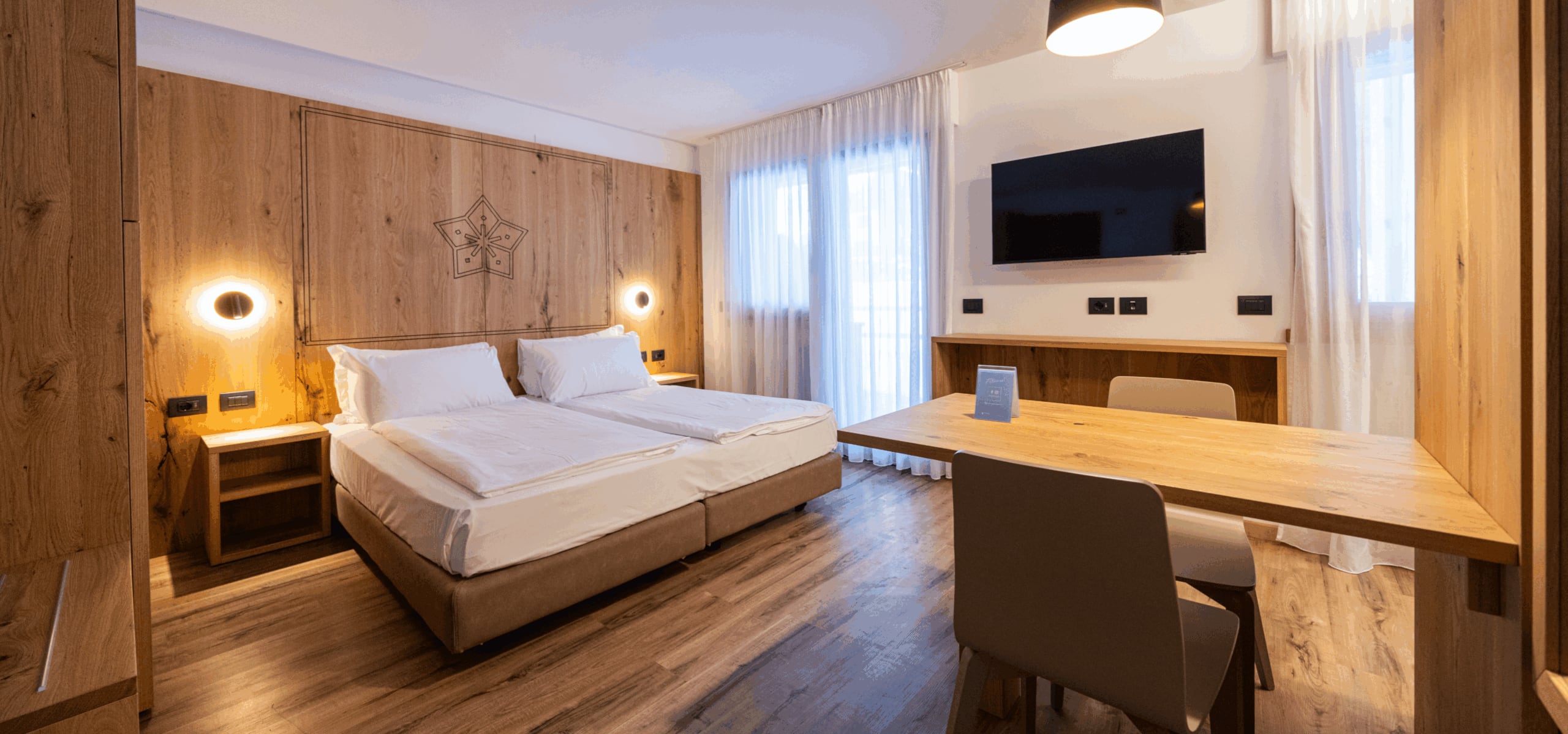 Camera d'albergo con due letti singoli affiancati, testiera in legno con decoro, lampade rotonde, scrivania e TV