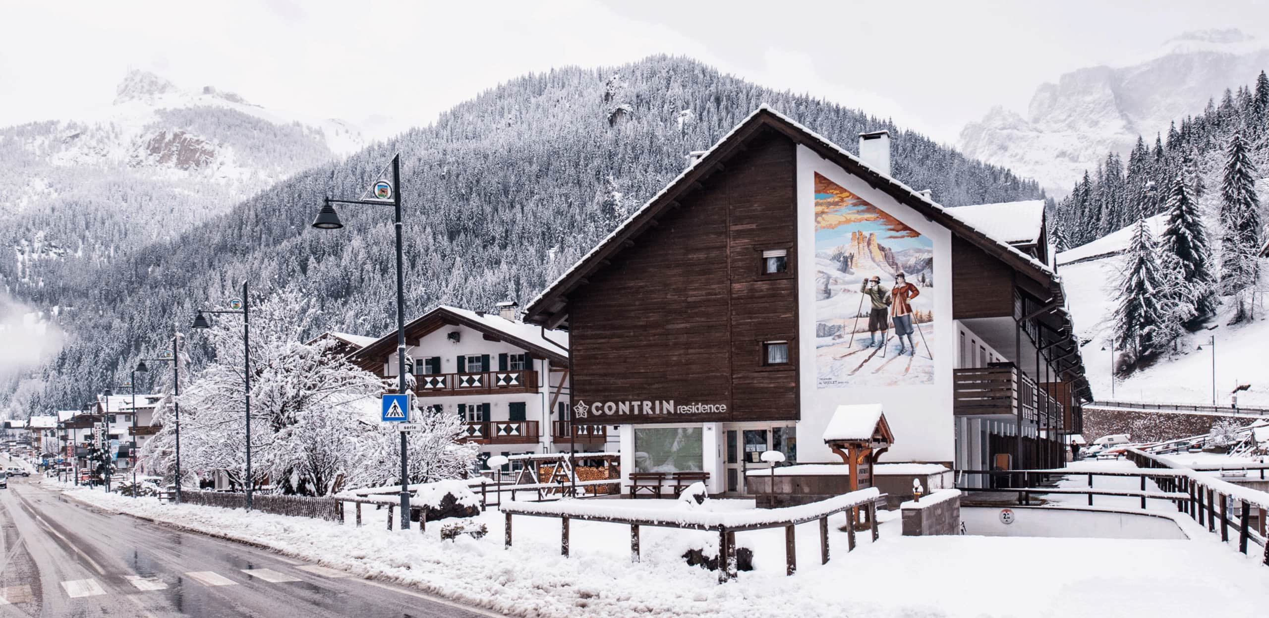 Edificio Contrin residence in paese innevato con murale di sciatori, strada e montagne boschive sullo sfondo