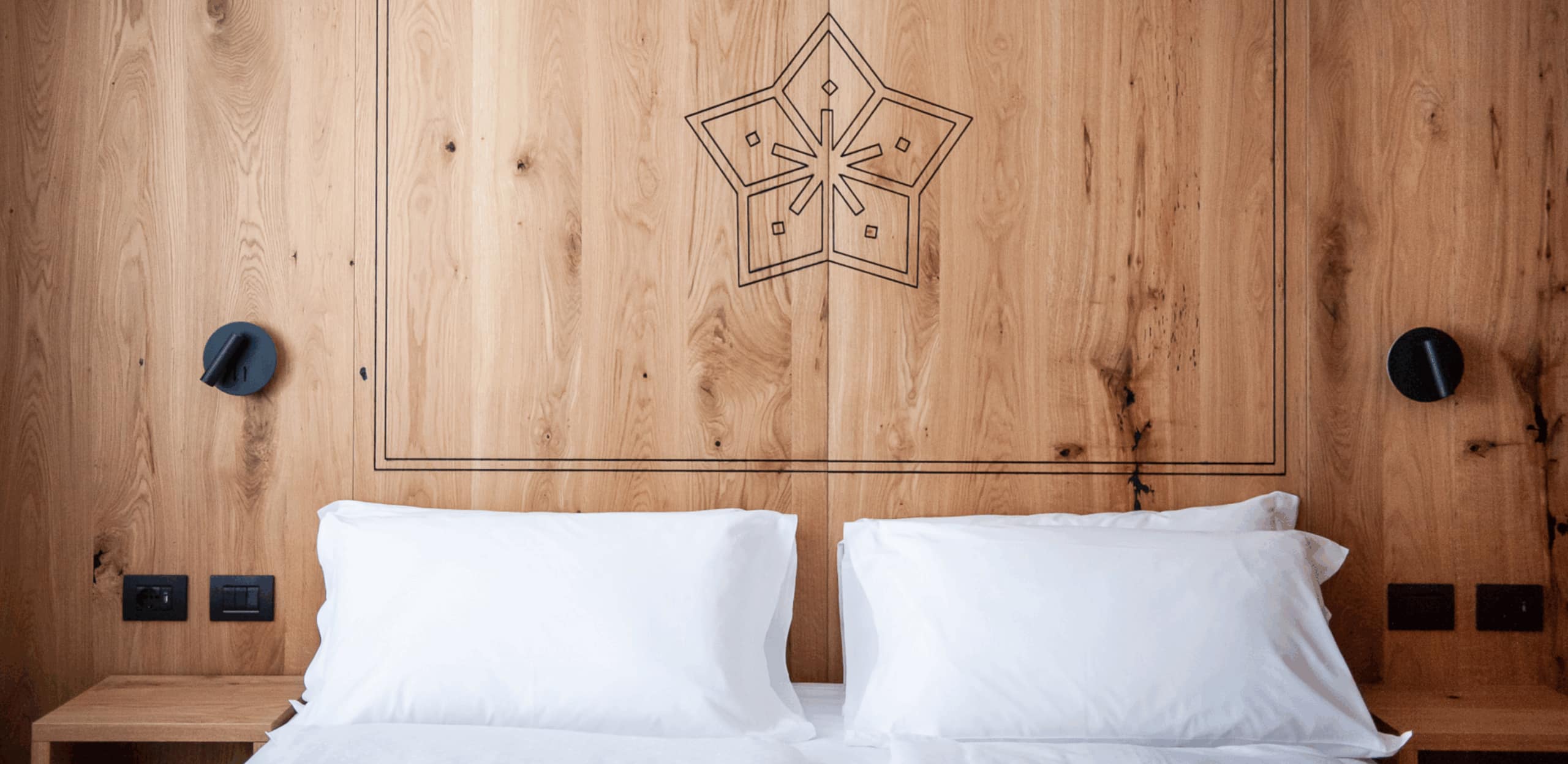Letto con due cuscini bianchi, testata in legno con motivo a stella, lampade e prese nere.