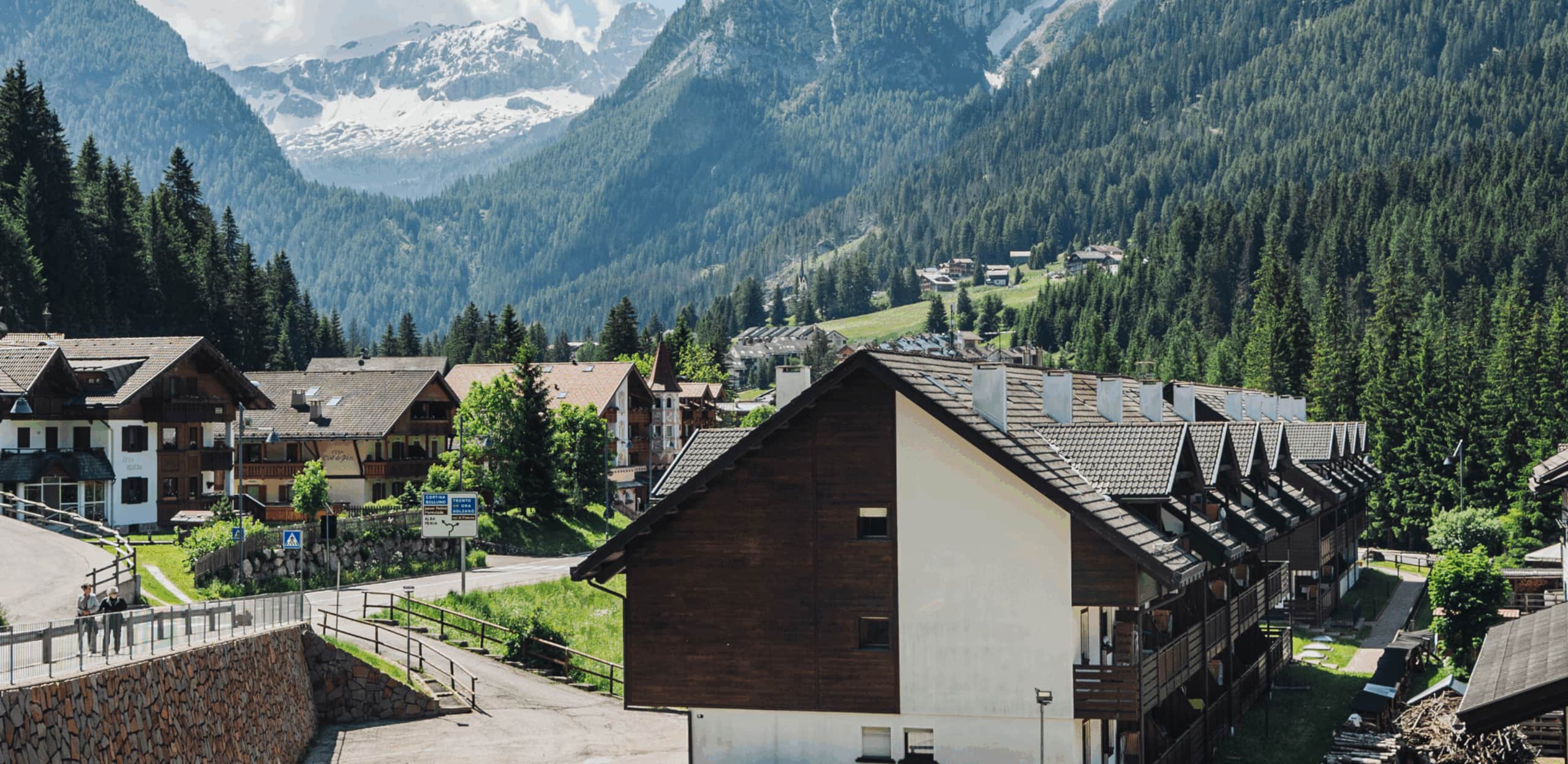 Fila di chalet in legno lungo una strada di montagna, boschi e cime innevate sullo sfondo; due persone a piedi.